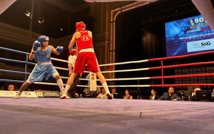 Zwei Boxer im roten und blauen Dress kämpfen im Ring beim 50. Chemiepokal in Halle, im Hintergrund ist das S&G Sponsorenlogo auf der Leinwand sichtbar.