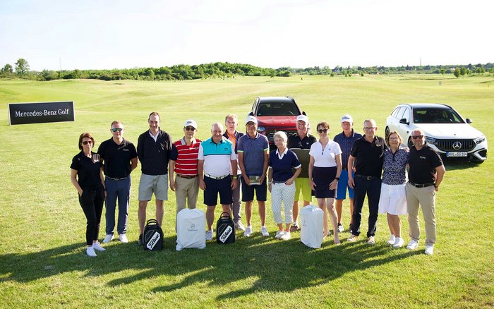 Gruppenfoto der Teilnehmerinnen und Teilnehmer des S&G Mercedes-Benz Golfturniers im Golfpark Hufeisensee auf dem Golfplatz, mit Mercedes-Benz Fahrzeugen im Hintergrund.