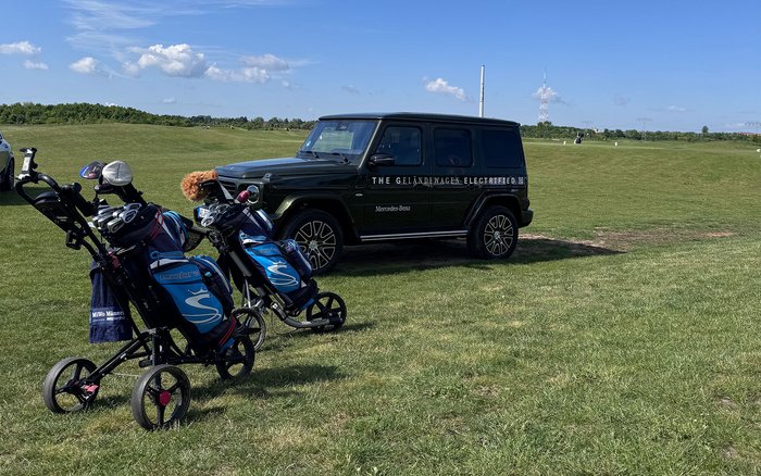 Zwei Golfbags auf Caddys vor einer Mercedes-Benz G-Klasse mit S&G Beschriftung auf einer weitläufigen Wiese im Golfpark Hufeisensee bei sonnigem Wetter