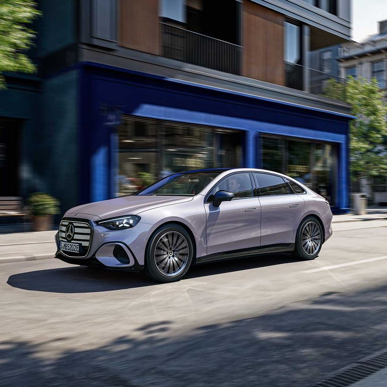 Silberne vollelektrische Mercedes-Benz C-Klasse fährt bei Tageslicht durch eine moderne Innenstadtstraße vor zeitgemäßer Architektur.
