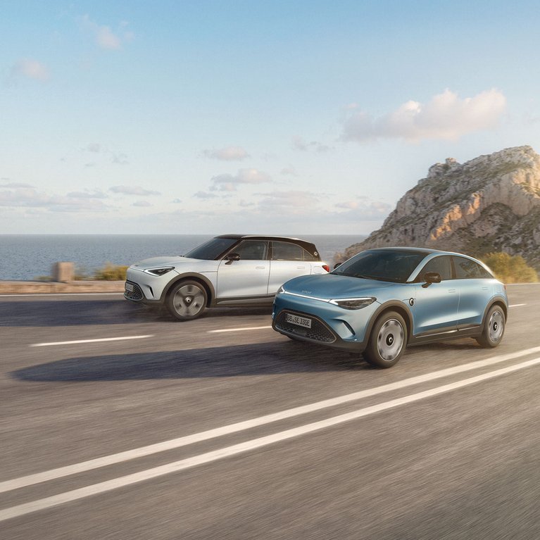 Zwei smart Fahrzeuge in der TUI Edition fahren an einer sonnigen Küstenstraße entlang – Symbol für Freiheit, Reisen und das exklusive Sondermodell von smart.