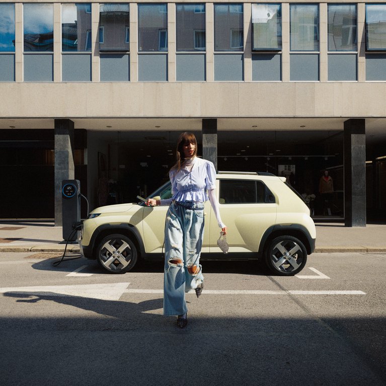 Promotionsbild für für Leasingangebote elektrischer Hyundai Fahrzeugmodelle. Eine Frau läuft vor dem Hyundai INSTER, der seitlich vor einem Gebäude geparkt ist.