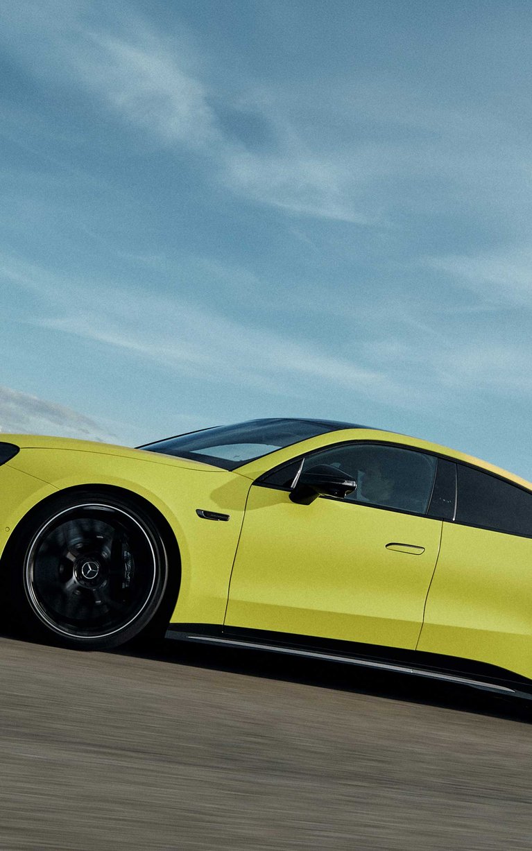 Gelbes Mercedes-AMG GT 4-Türer Coupé fährt dynamisch auf einer Rennstrecke vor blauem Himmel.