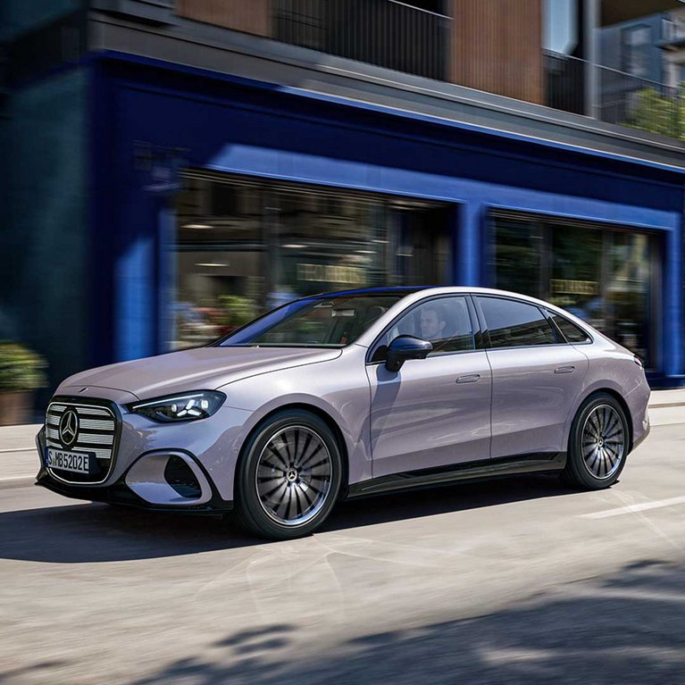 Silberne vollelektrische Mercedes-Benz C-Klasse fährt bei Tageslicht durch eine moderne Innenstadtstraße vor zeitgemäßer Architektur.