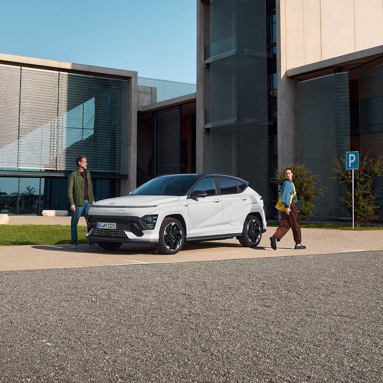 Weißer Hyundai KONA Elektro vor einem modernen Gebäude mit Personen im Außenbereich