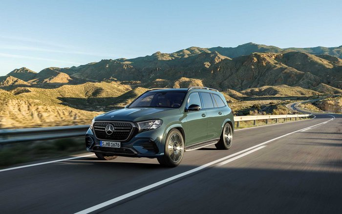 Neuer Mercedes-Benz GLS in Dunkelgrün fährt auf einer Straße entlang einer bergigen Küstenlandschaft bei Tageslicht.