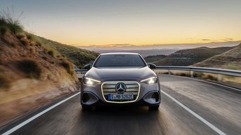 Mercedes-Benz Elektrofahrzeug auf einer kurvenreichen Straße in einer bergigen Landschaft bei Sonnenuntergang, Frontansicht während der Fahrt.