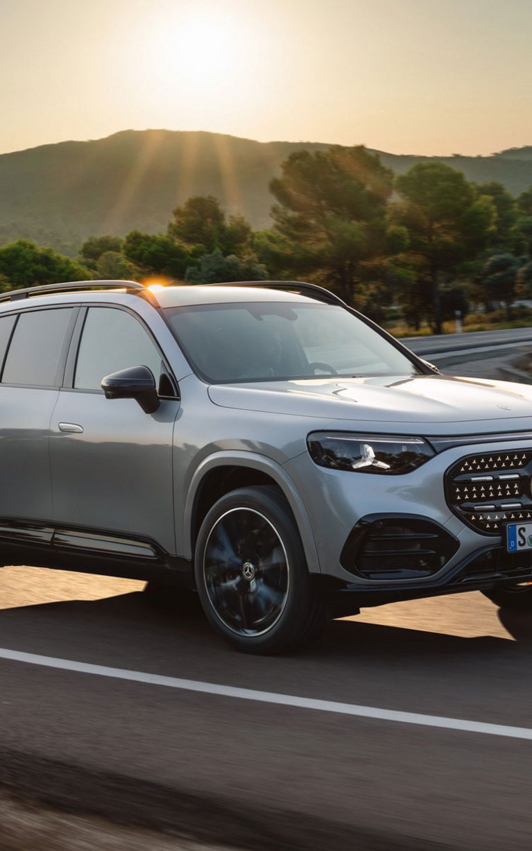 Mercedes-Benz GLB mit EQ Technologie fährt auf einer kurvigen Landstraße bei Sonnenuntergang durch eine grüne Landschaft; modernes SUV in hellem Farbton.