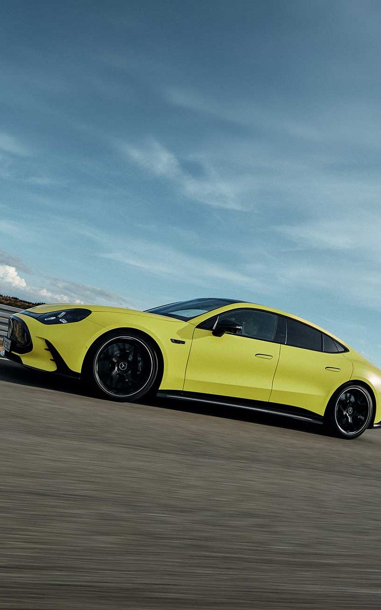 Gelbes Mercedes-AMG GT 4-Türer Coupé fährt dynamisch auf einer Rennstrecke vor blauem Himmel.
